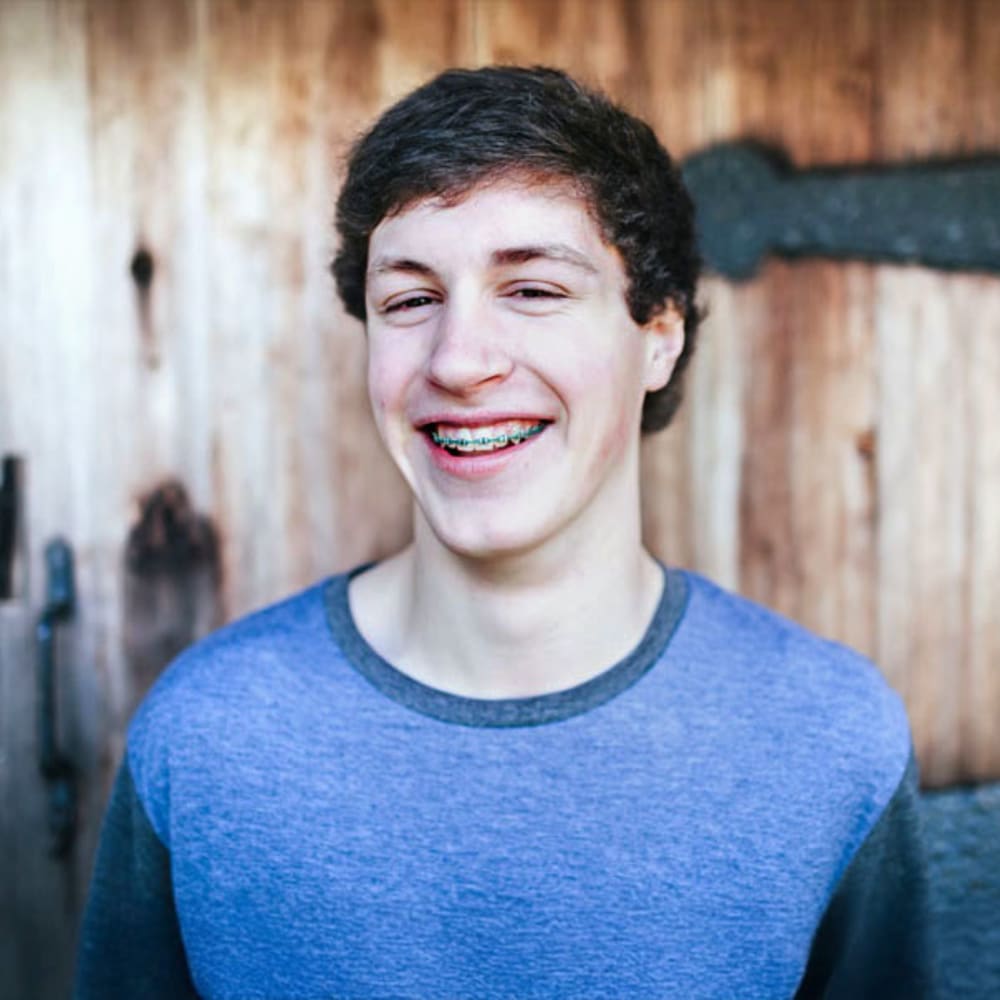 A white man in his late teens/early 20s is wearing a blue shirt and smiling, revealing blue braces.