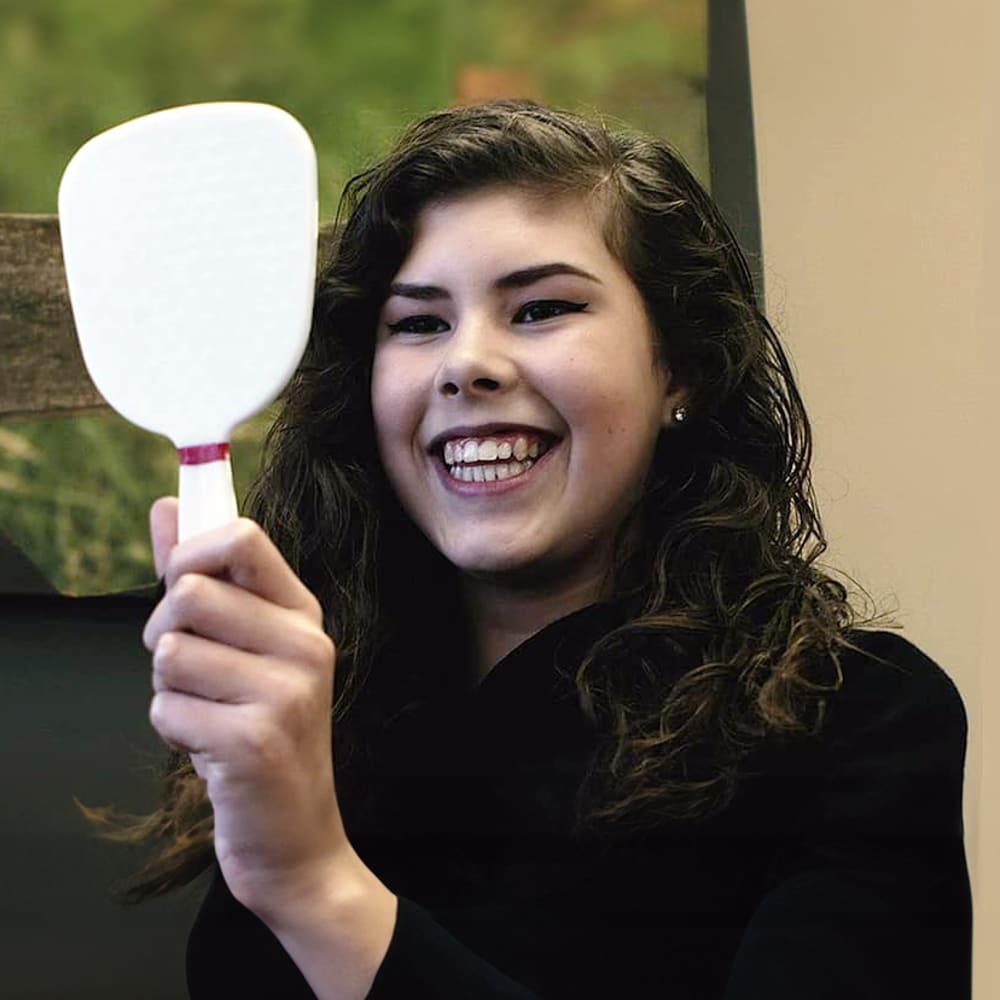 A young adult woman smiles into a handheld mirror.