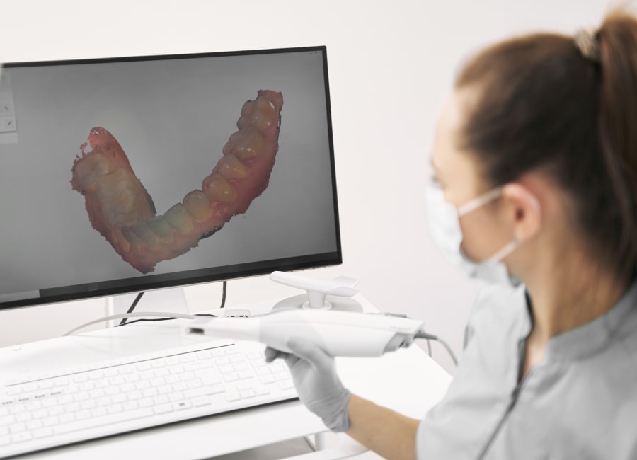 A female dental professional holds an intraoral scanner while looking at a monitor in the background which shows an image of the teeth and gums.
