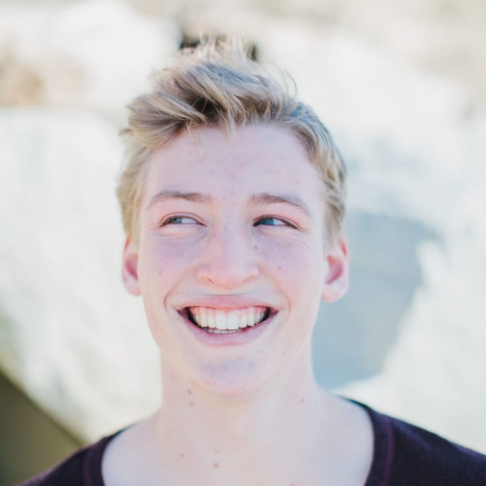 A teenage boy with blonde hair and blue eyes is smiling and looking to the left.