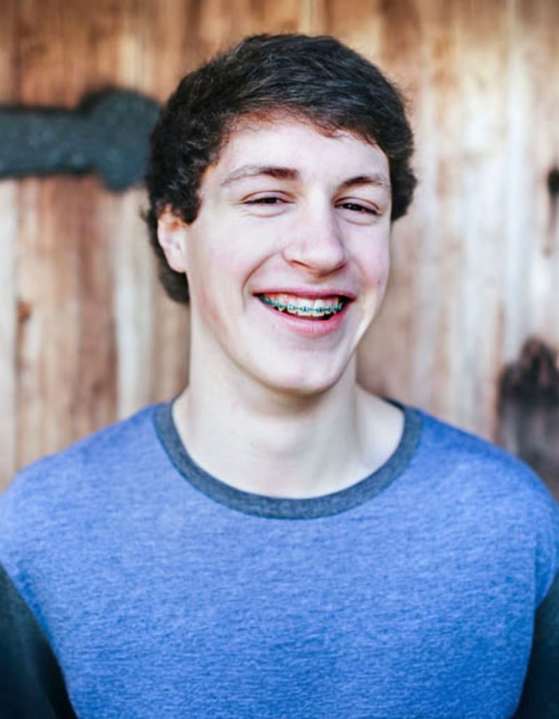Teenage boy standing outside in front of a wooden door, smiling.
