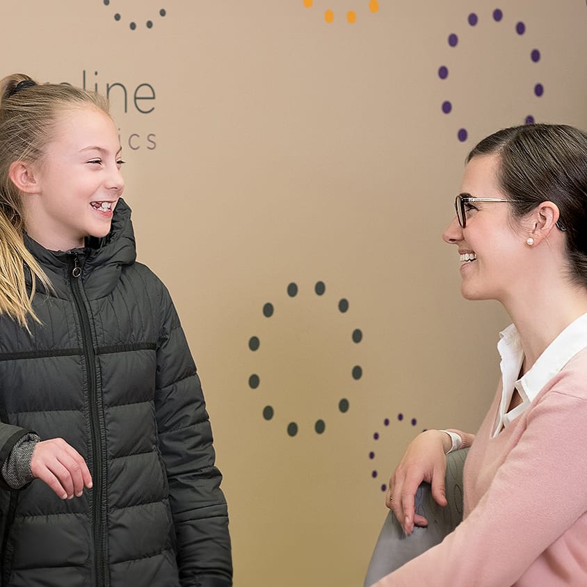 A young girl smiles while standing in front of a Shoreline Orthodontics wall. She is talking to Dr. McFadden.